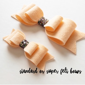 Może przedstawiać: Dwa filcowe kokardki w kolorze brzoskwiniowym z błyszczącym środkiem. Tekst "standard or super felt bows" jest napisany pod kokardkami.
