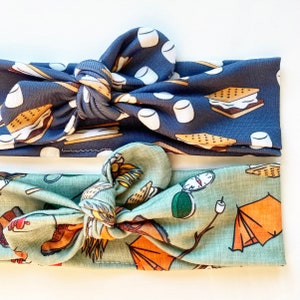 May include: Two fabric headbands with a knotted design. The top headband is navy blue with a pattern of marshmallows, graham crackers, and chocolate squares. The bottom headband is light green with a pattern of camping gear, including a tent, compass, and hiking boots.