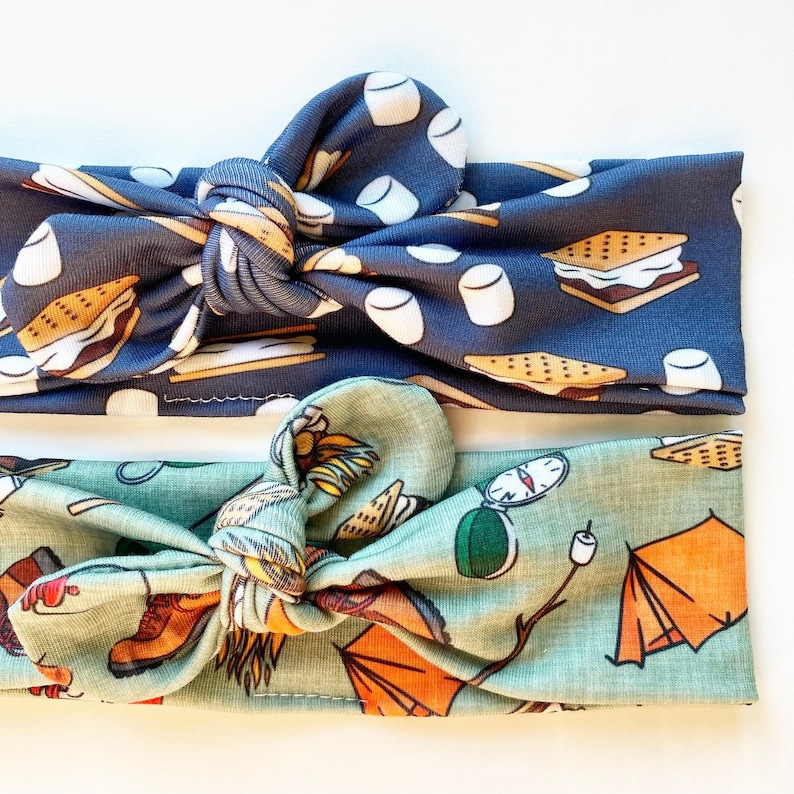 May include: Two fabric headbands with a knotted design. The top headband is navy blue with a pattern of marshmallows, graham crackers, and chocolate squares. The bottom headband is light green with a pattern of camping gear, including a tent, compass, and hiking boots.