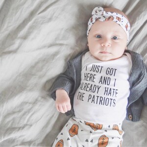 May include: A baby wearing a white onesie with the text "I just got here and I already hate the Patriots" and a gray cardigan. The baby is also wearing white pants with a football pattern and a white headband with a football pattern.