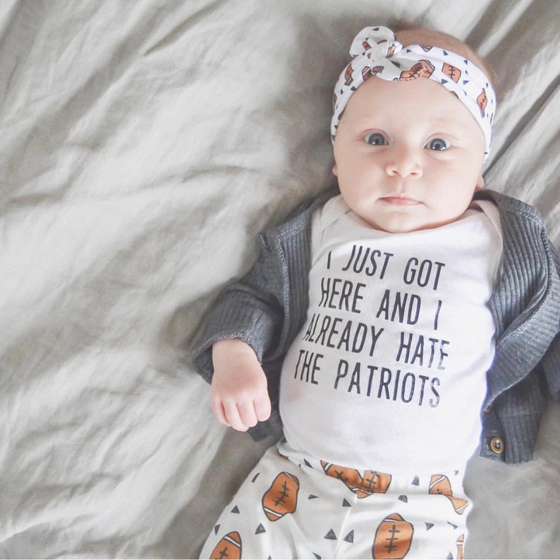May include: A baby wearing a white onesie with the text "I just got here and I already hate the Patriots" and a gray cardigan. The baby is also wearing white pants with a football pattern and a white headband with a football pattern.