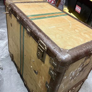 May include: A vintage wooden trunk with brown metal trim and hardware. The trunk has green and orange stripes on the top. It features metal corner protectors and multiple latches. The trunk is a light brown colour.