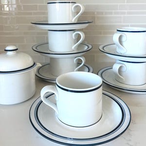 May include: White ceramic teacups and saucers with a navy blue trim. The set includes a teapot with a lid and several stacked cups and saucers. The cups have curved handles.