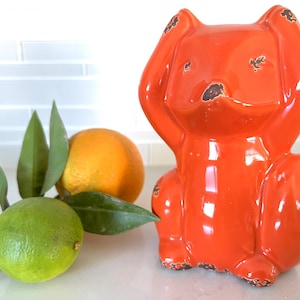 May include: A bright orange ceramic fox ornament with a distressed finish, with its paws raised. A lime, an orange, and green leaves are placed beside it, suggesting a decorative or kitchen theme.
