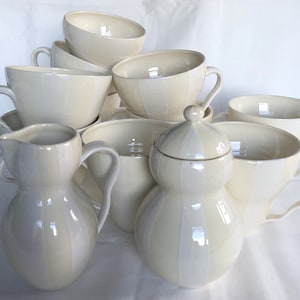 Può includere: Una collezione di stoviglie in ceramica color crema, tra cui tazze da tè, una lattiera e una zuccheriera con coperchio. I pezzi presentano un design a righe verticali. Le tazze da tè hanno grandi manici. La zuccheriera ha una forma arrotondata.