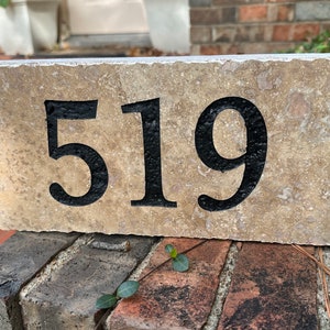 May include: A rectangular address stone with the number 519 in black, set against a beige, textured background. The stone is resting on a brick structure, with greenery visible in the background.