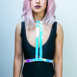 May include: A woman with pink hair is wearing a black swimsuit with a holographic iridescent chest harness. The harness has two straps that cross in the front and connect at the back.