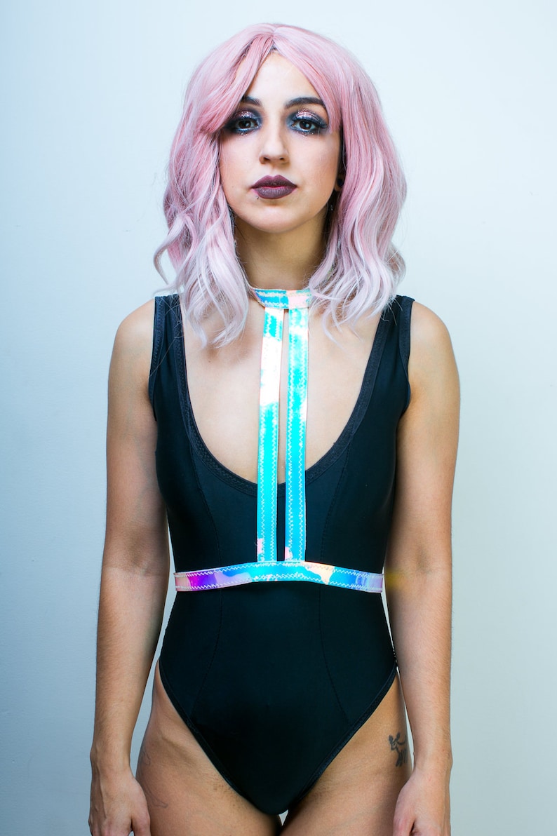 May include: A woman with pink hair is wearing a black swimsuit with a holographic iridescent chest harness. The harness has two straps that cross in the front and connect at the back.