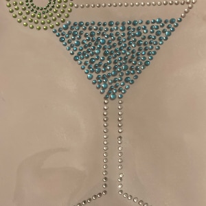 May include: A cocktail glass design made of sparkling rhinestones. The glass is filled with blue rhinestones, with a lime wedge made of green rhinestones. The stem and rim are outlined with clear rhinestones.