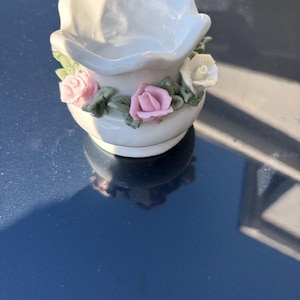 May include: A small, white ceramic dish with a scalloped rim. The dish is decorated with pink and white roses and green leaves around the base. The dish is likely a decorative item.