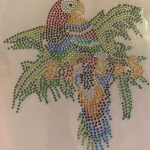 May include: A colorful parrot design made of sparkling rhinestones. The bird features red, blue, yellow, green, and black gems arranged on a white mesh background. The parrot's wings are spread, and the design is detailed.
