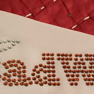 May include: A white fabric with the word "Bewitched" spelled out in orange rhinestones. A witch hat made of silver and orange rhinestones is above the word. The background is a red quilted fabric.