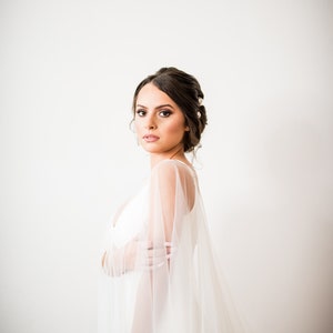 May include: A woman in a white wedding dress with a sheer, flowing cape. Her dark hair is styled up with pearl accents. She is looking at the camera. The background is a plain white wall.