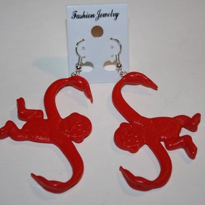May include: A pair of red monkey earrings. The monkeys are in a playful pose with their arms and legs outstretched. The earrings are made of plastic and have silver hooks.