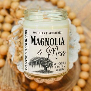 May include: A clear glass jar candle filled with off-white soy wax. The label reads Southern E-Scentuals, Magnolia & Moss, Soft Scent, Shaded Blooms, 7 oz / 198 g, 50 hours, and features a black and white illustration of a tree.