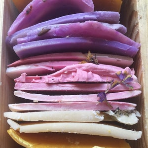 May include: A stack of handmade soap bars in various shades of purple, pink, and white. The soap bars are layered on top of each other and have dried flowers and herbs embedded in them.