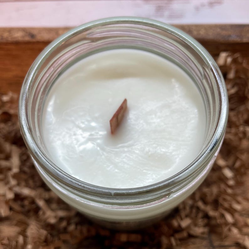 May include: A clear glass jar containing white candle wax with a single flat wooden wick centered in the middle. The surface of the wax is smooth and opaque, filling the circular opening of the glass container completely from a top-down view.