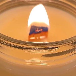 May include: A lit candle in a clear glass jar with a wooden wick. The flame is bright and centered, casting a warm glow over the smooth, light-colored wax surface. The wooden wick is partially charred and dark at the base, surrounded by wax.