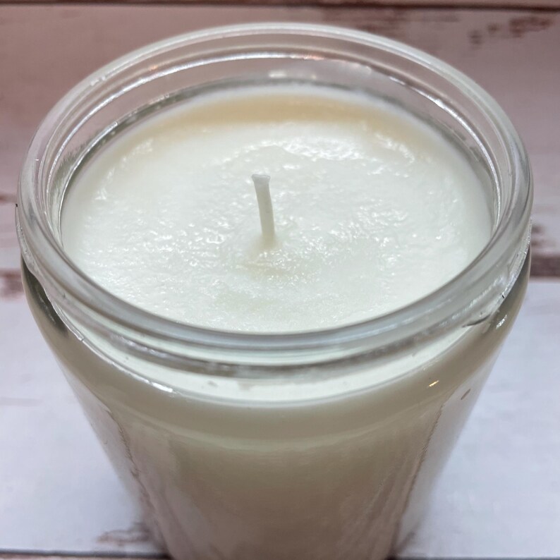 May include: A white candle in a clear glass jar with a single centered wick. The wax surface is smooth and opaque, filling the cylindrical container to the brim. The glass jar features a thick rim and transparent walls, showing the white wax inside.