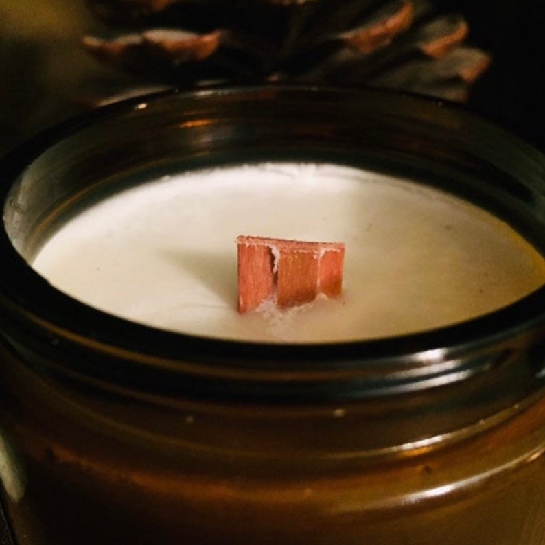 May include: A candle in a dark amber glass jar with smooth white wax and a flat, rectangular wooden wick centered in the middle. The wick is a light reddish-brown color and stands upright from the surface of the wax, which fills the container.