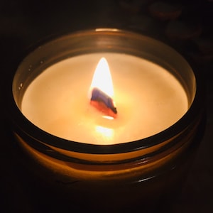 May include: A lit candle in a dark amber glass jar with a visible wooden wick burning in the center. The wax is off-white and smooth, filling the cylindrical container. The flame is bright and orange, casting a warm glow over the wax surface.