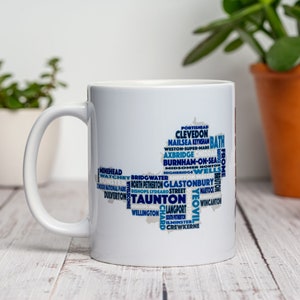 May include: White ceramic mug with a blue and white design featuring the names of towns and cities in Somerset, England. The design is in the shape of the county of Somerset.