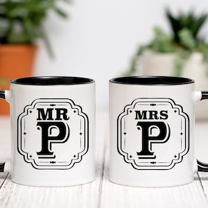 May include: Two white ceramic mugs with black rims and handles. The mugs feature a black and white design with the words "MR P" and "MRS P" in a vintage style font.