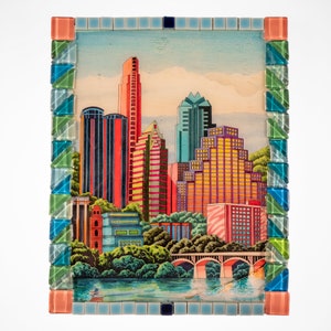 May include: A colorful mosaic framed artwork depicting a cityscape with tall buildings in various shades of pink, red, yellow, blue, and green. The buildings are set against a blue sky with white clouds. A bridge is visible in the foreground.
