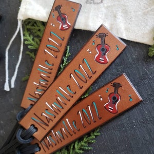 May include: Three brown leather bookmarks with black leather straps. Each bookmark has a hand-painted guitar and the text "Nashville 2022" in white.