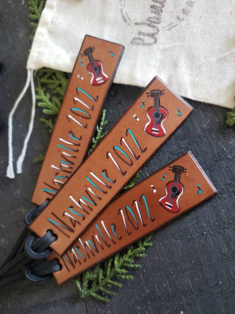 May include: Three brown leather bookmarks with black leather straps. Each bookmark has a hand-painted guitar and the text "Nashville 2022" in white.