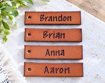 Peut inclure: Quatre étiquettes rectangulaires en cuir beige avec des bords marron foncé, chacune dotée d'un trou circulaire sur le côté gauche et d'un nom gravé à l'encre noire. Les noms Brandon, Brian, Anna et Aaron sont centrés sur chaque pièce.