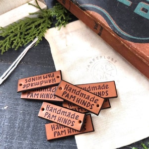 May include: A collection of brown leather tags with the words "Handmade PAM HINDS" printed in black. The tags are rectangular with rounded corners and two holes. A vintage book and a canvas bag are in the background.