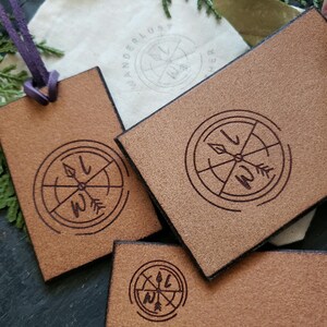 May include: Close-up of three brown leather tags with a compass design engraved on each. The text "BACKSDE OF TAGS" and "(CANNOT BE ENGRAVED, THE GRAIN DOESN'T ENGRAVE CLEARLY ON THIS SIDE, USED FOR LOGO)" is printed on the bottom of the image.