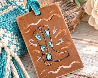 May include: A rectangular tan leather tag featuring a hand-painted illustration of a cowboy boot in brown, white, and turquoise. The tag is suspended by a bright turquoise leather cord and displays decorative wavy lines in white and orange.