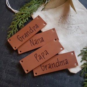 May include: Four rectangular tan leather tags with pre-punched holes at each corner. The tags are inscribed with the black text Grandpa, Nana, Papa, and Grandma. They are arranged on a cream-coloured fabric pouch featuring a circular logo.