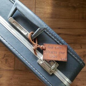 May include: A blue suitcase with a leather luggage tag that says "Your vibe attracts your tribe".