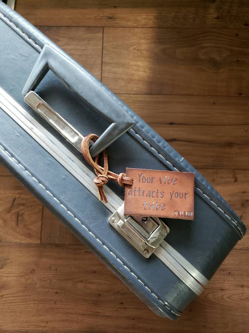 May include: A blue suitcase with a leather luggage tag that says "Your vibe attracts your tribe".