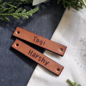 May include: Two brown leather labels with the names "Yogi" and "Harshy" engraved on them. The labels are on a white fabric surface.