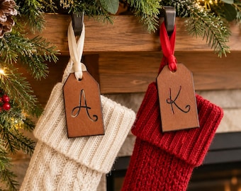 以下が含まれることがあります： クリーム色と赤色のニット製クリスマスストッキングが2つ、暗い色の金属製フックに掛けられています。それぞれのストッキングには、黒い文字（AとK）が刻印された長方形の茶色のレザータグがリボンで取り付けられています。