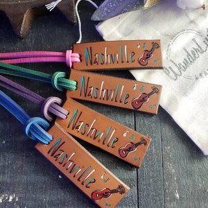 May include: Four brown leather keychains with the word "Nashville" written in black and a small painted guitar with musical notes. The keychains are attached to colorful cords.