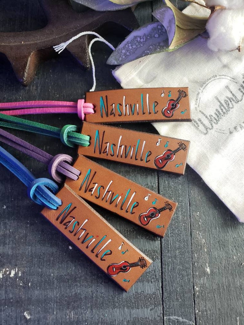 May include: Four brown leather keychains with the word "Nashville" written in black and a small painted guitar with musical notes. The keychains are attached to colorful cords.