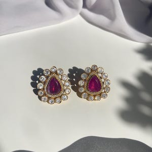 May include: A pair of gold earrings featuring a teardrop-shaped purple gemstone surrounded by smaller clear gemstones.