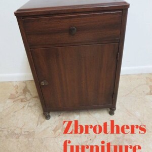 May include: A dark wood nightstand with a single drawer and a cabinet door. The nightstand has brass hardware and sits on four small feet.