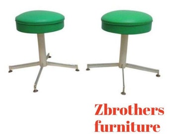 May include: Two green upholstered stools with a silver metal base and three legs. The stools are on a white background.