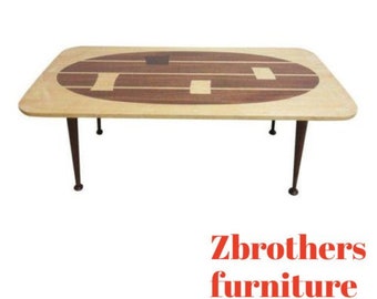 May include: A mid-century modern coffee table with a light wood top and a dark wood inlay design. The table has four black metal legs.