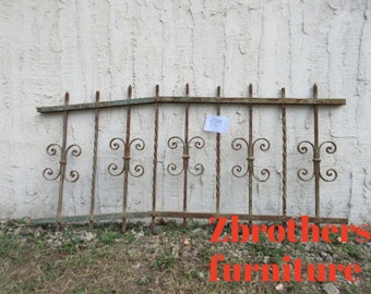 May include: A rusty metal fence panel with a curved top and decorative scrollwork. The fence is made of iron bars with a twisted design. The fence is against a white wall.