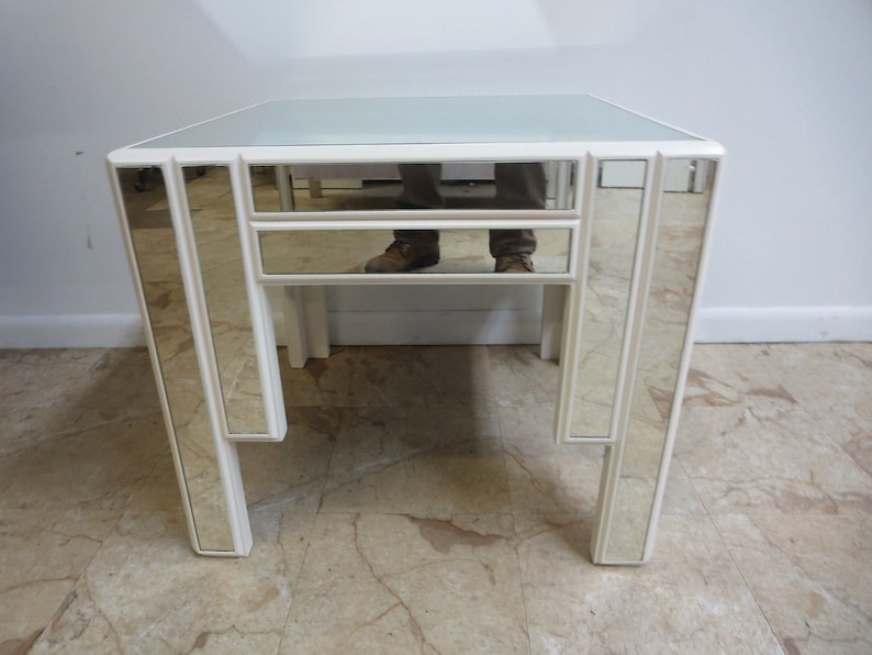 May include: A white mirrored end table with a geometric design. The table has a glass top and a single drawer.