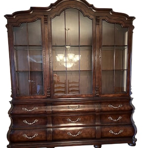 May include: A large, dark brown wooden hutch with glass-paned doors and drawers. The hutch has a curved design and ornate hardware. The glass doors reflect a chandelier and chairs. A classic piece of furniture.