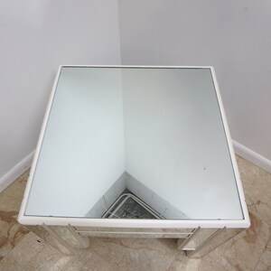 May include: A white mirrored end table with a square top and a beveled edge. The table has a mirrored surface and a white frame.