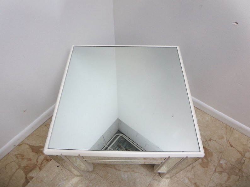 May include: A white mirrored end table with a square top and a beveled edge. The table has a mirrored surface and a white frame.